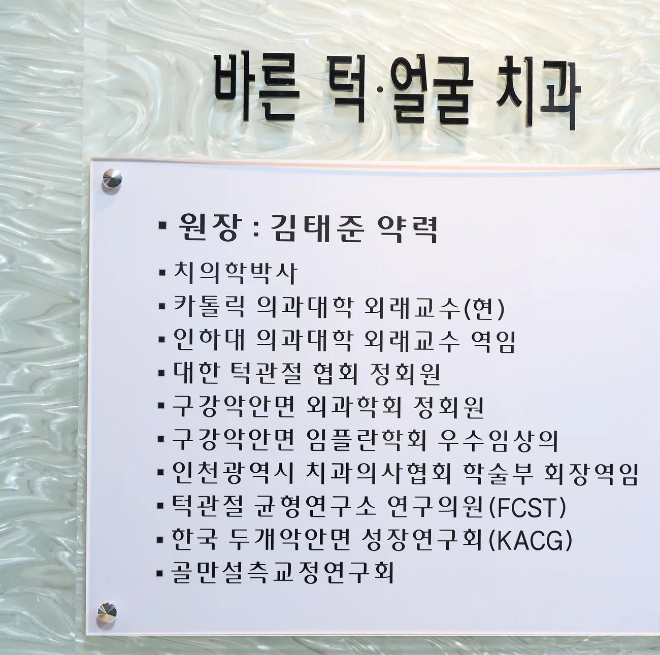 바른턱얼굴치과의원 입구에 게시된 김태준 대표원장 약력 명판 — 치의학박사, 카톨릭 의과대학 외래교수(현), 인하대 의과대학 외래교수 역임, 대한 턱관절 협회 정회원, 구강악안면 외과학회 정회원, 구강악안면 임플란트학회 우수임상의, 인천광역시 치과의사협회 학술부 회장역임, 턱관절 균형연구소 연구의원(FCST), 한국 두개악안면 성장연구회(KACG), 골만설측교정연구회